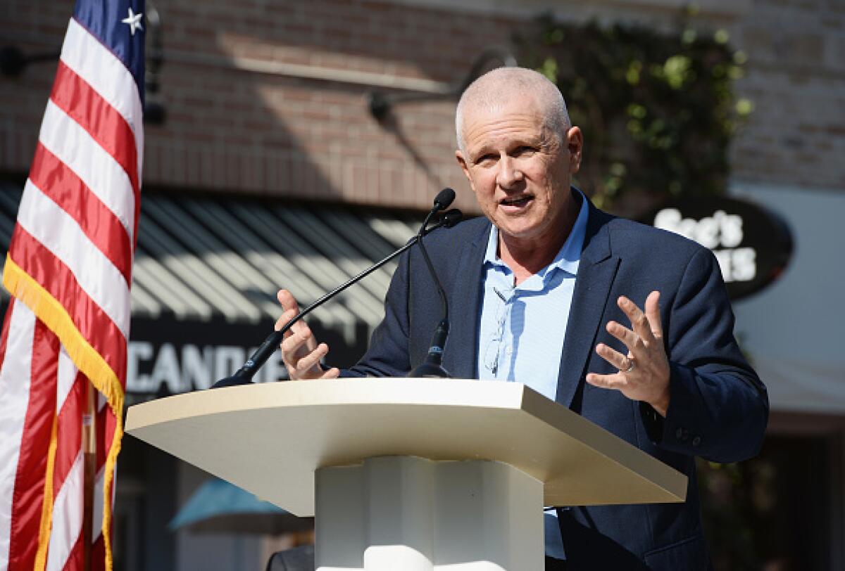 A man in a suit jacket stands outside at a lectern next to a flag speaking into a microphone.