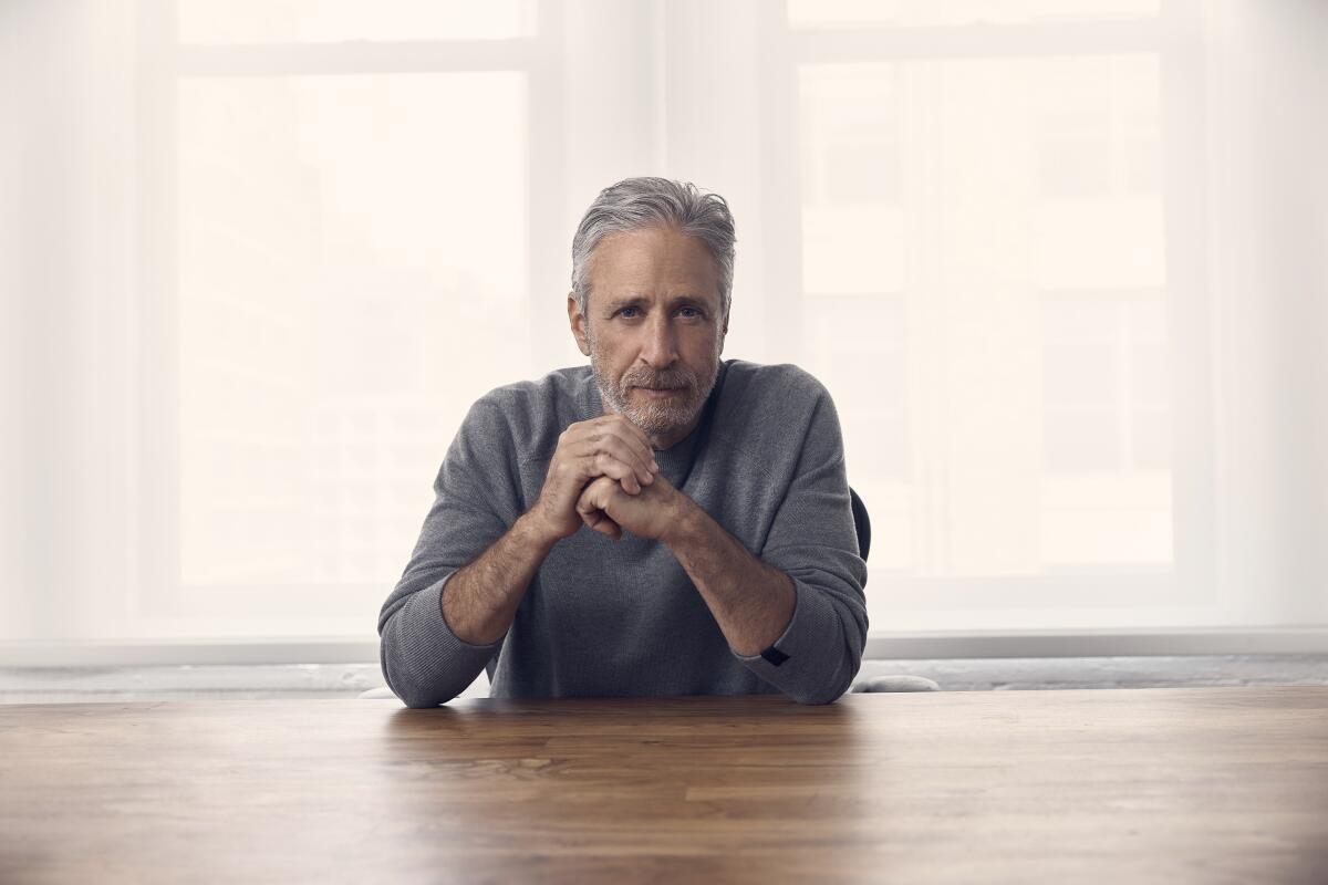 A bearded man in a gray sweater sits with his elbows on a desk and his hands near his chin.