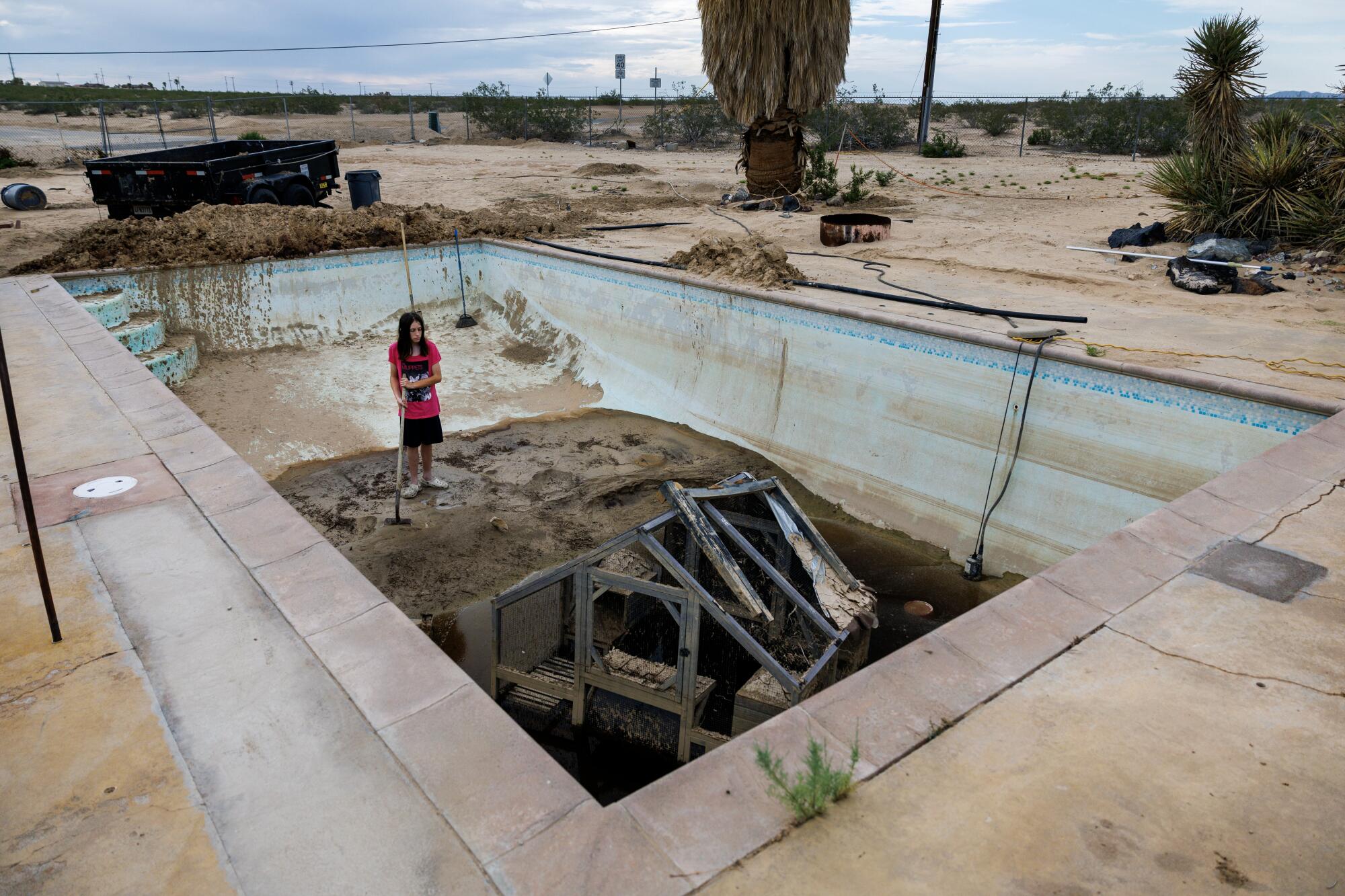 A person in a red shirt and dark shorts stand atop a pile of mud in an empty swimming pool 