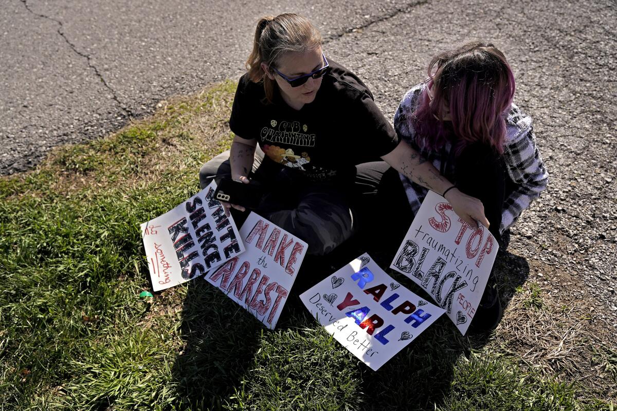 A mother and daughter sit on the ground with protest signs reading "Make the arrest" and "White silence kills." 