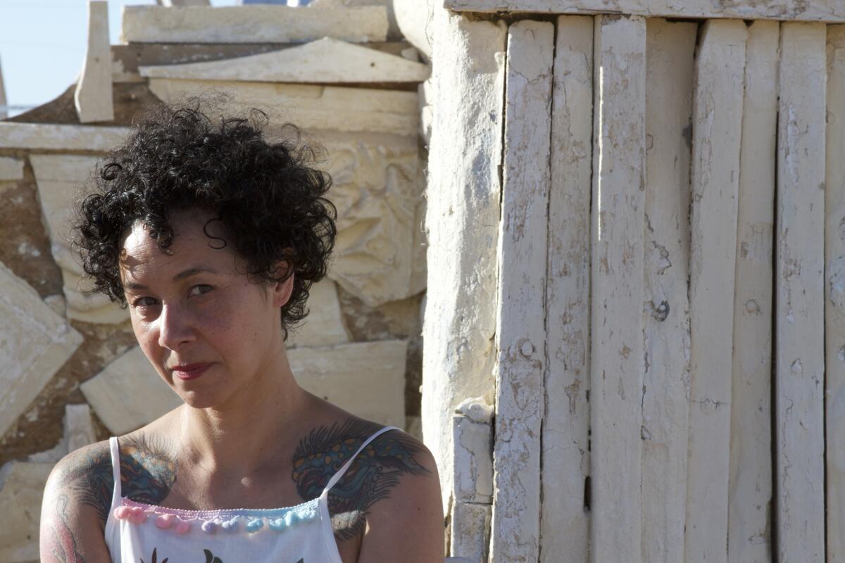 A woman from the shoulders up outside a stone, desert-y house 