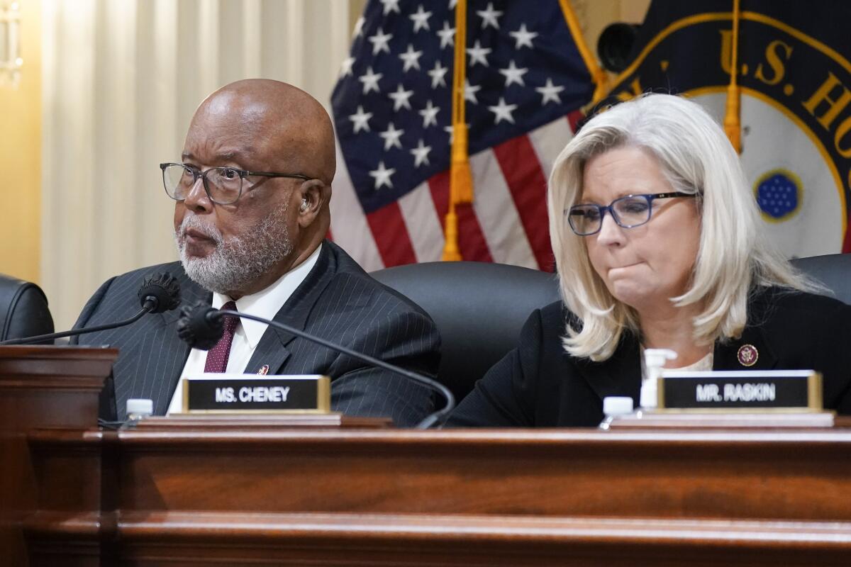 A man with a shaved head, in suit and tie, and a woman with blond hair, both wearing glasses, are seated in front of flags 