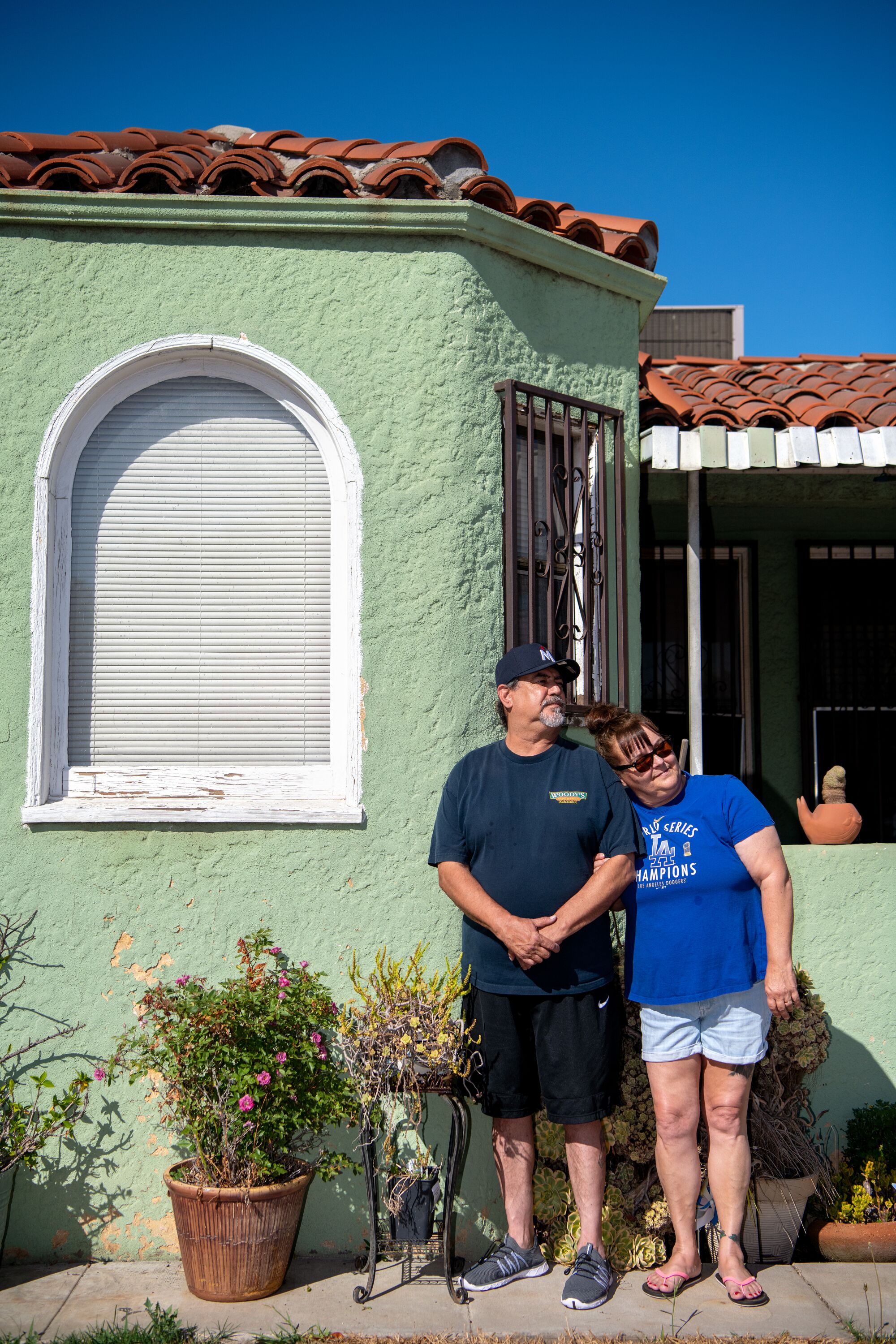 A man and woman stand outside a stucco-walled house with a tile roof.