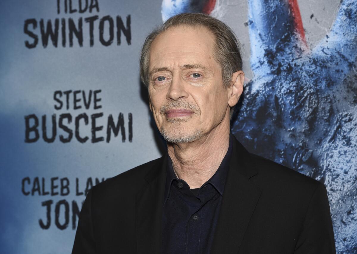 Steve Buscemi in a dark outfit posing against a blue background with black text