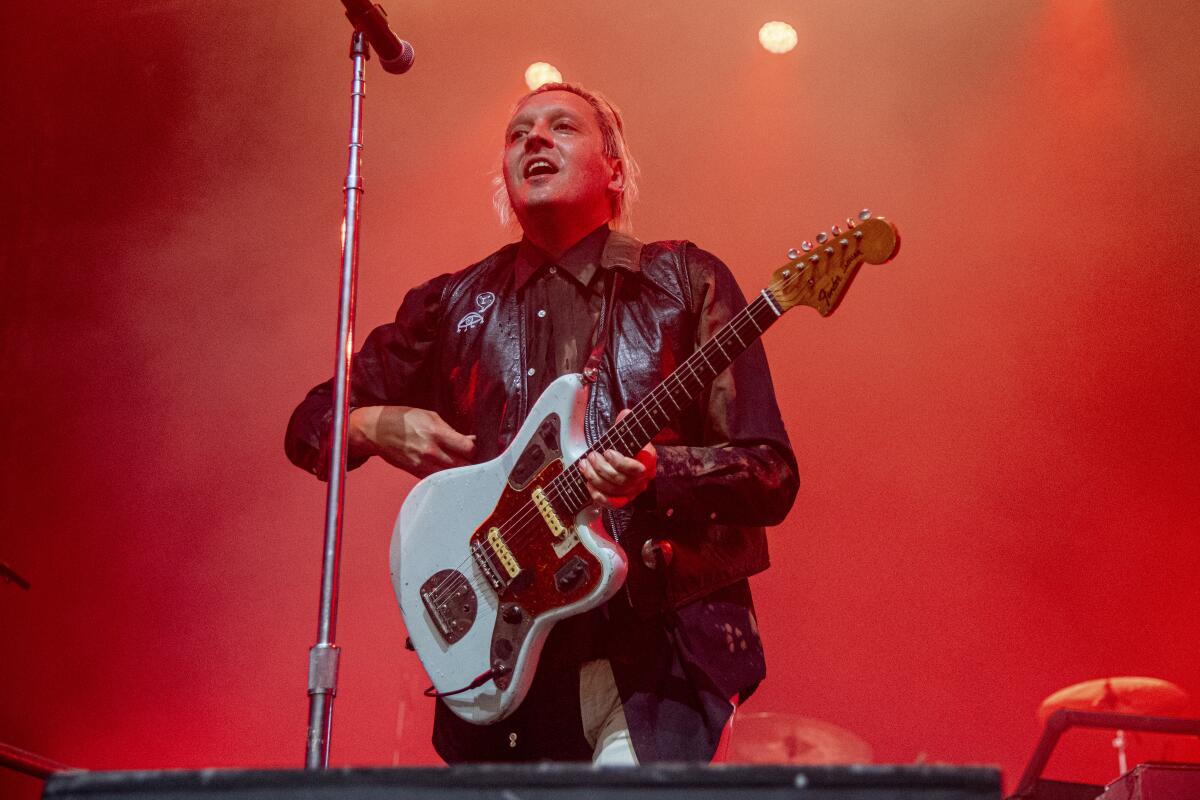 A man with platinum blond hair singing into a microphone and holding a guitar