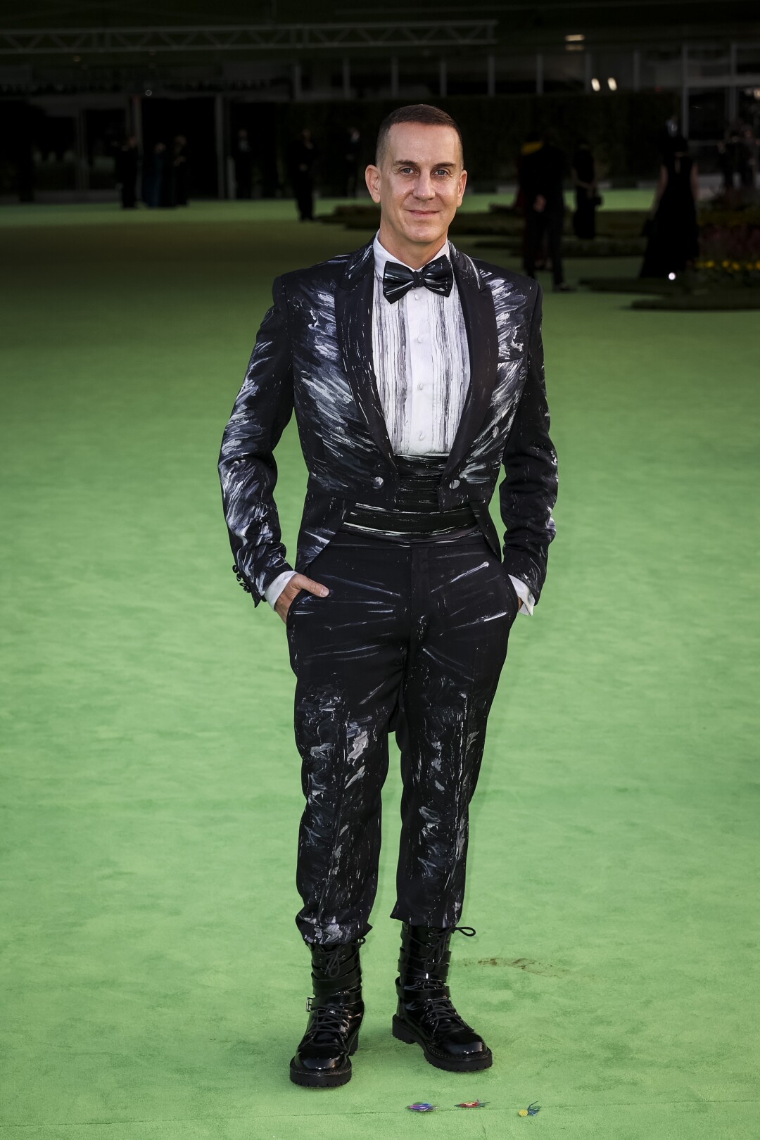 A man in a patterned black tuxedo and bowtie posing on a green carpet