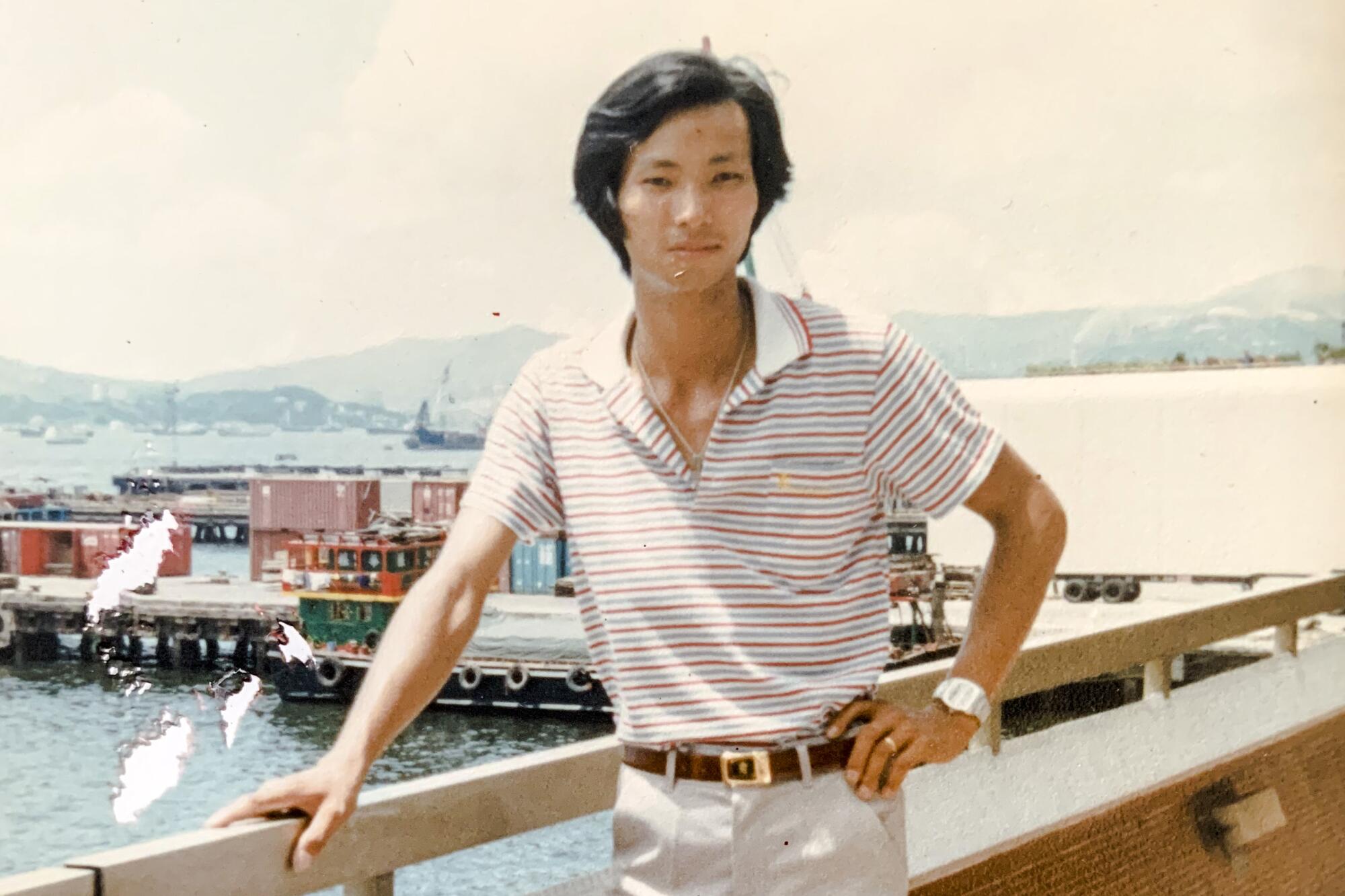 A man holds on to a wooden railing at a harbor while looking forward with a hand on his hip in an early 1980s photo