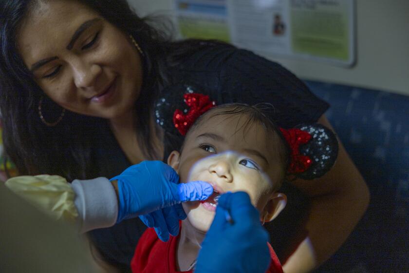 Long Beach, CA - October 12: Dental Hygienist Elizabeth Valdivia, left, cleans the teeth of Meliah Garcia, 2, as her mom, Carolina Garcia, of Lynwood, holds her at the Oral Health Education Center, an early intervention program that takes care of low-income children at Children's Dental Health Clinic at Children's Dental Health Clinic in Long Beach Thursday, Oct. 12, 2023. (Allen J. Schaben / Los Angeles Times)