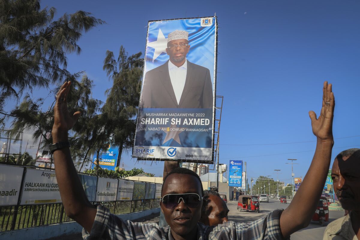 Supporters raise their hands near a campaign poster for former president and presidential candidate Sharif Sheikh Ahmed, on a street in Mogadishu, Somalia Tuesday, May 10, 2022. Somalia is set to hold its long-delayed presidential vote on Sunday, ending the convoluted electoral process that raised tensions in the country when the president's term expired last year without a successor in place. (AP Photo/Farah Abdi Warsameh)