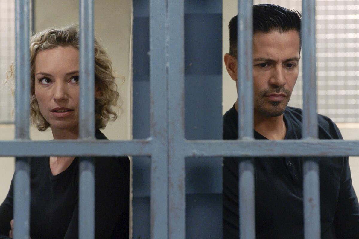 A woman and a man stand behind bars in adjoining jail cells.