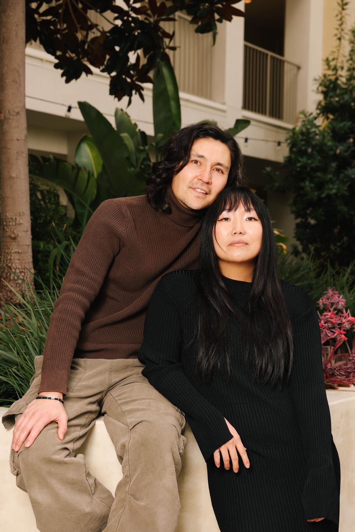 A man and a woman posing in front of shrubbery and the beige facade of an apartment building.