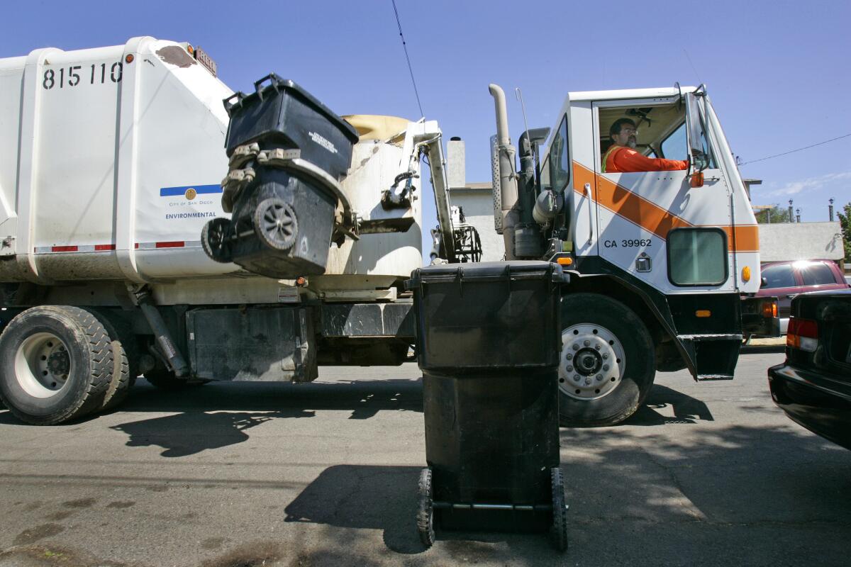A sanitation driver watches from his garbage truck window as the truck's hydraulic arm lifts a trash can
