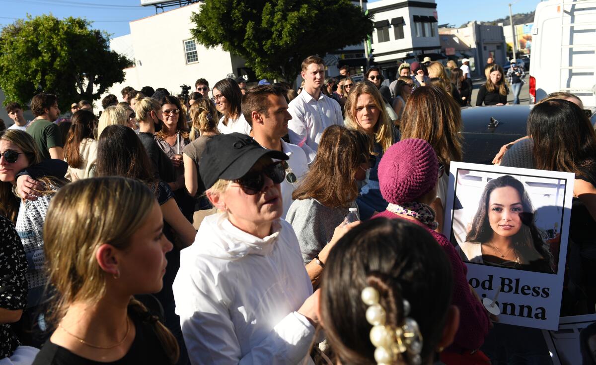 A crowd gathers outdoors. One person holds a woman's photo with the words "God Bless Breanna"