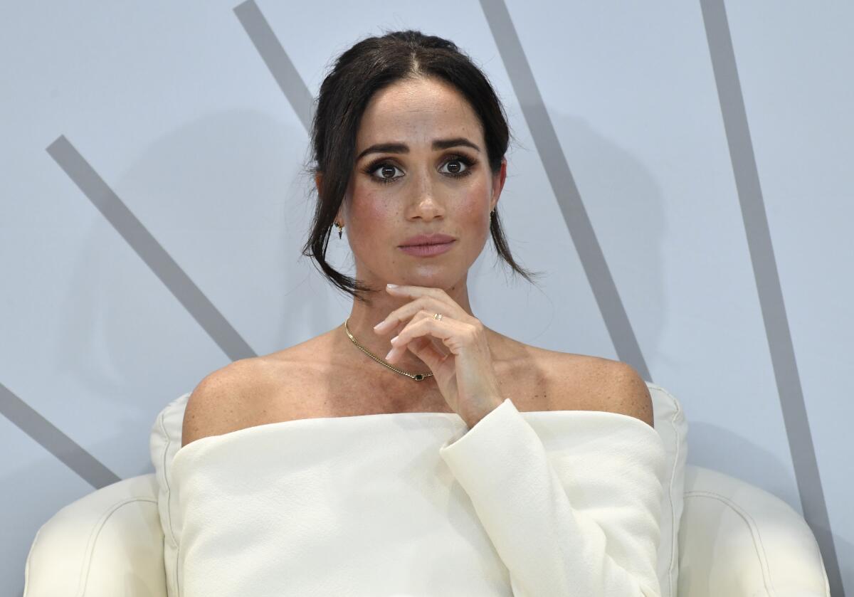 Meghan, Duchess of Sussex, sits in a white chair and stares pensively ahead while wearing a white outfit.