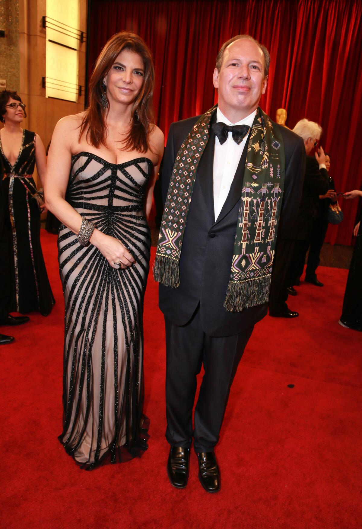 Dina De Luca Chartouni, in a black strapless dress, walks the red carpet with Hans Zimmer, in a black tuxedo and scarf.