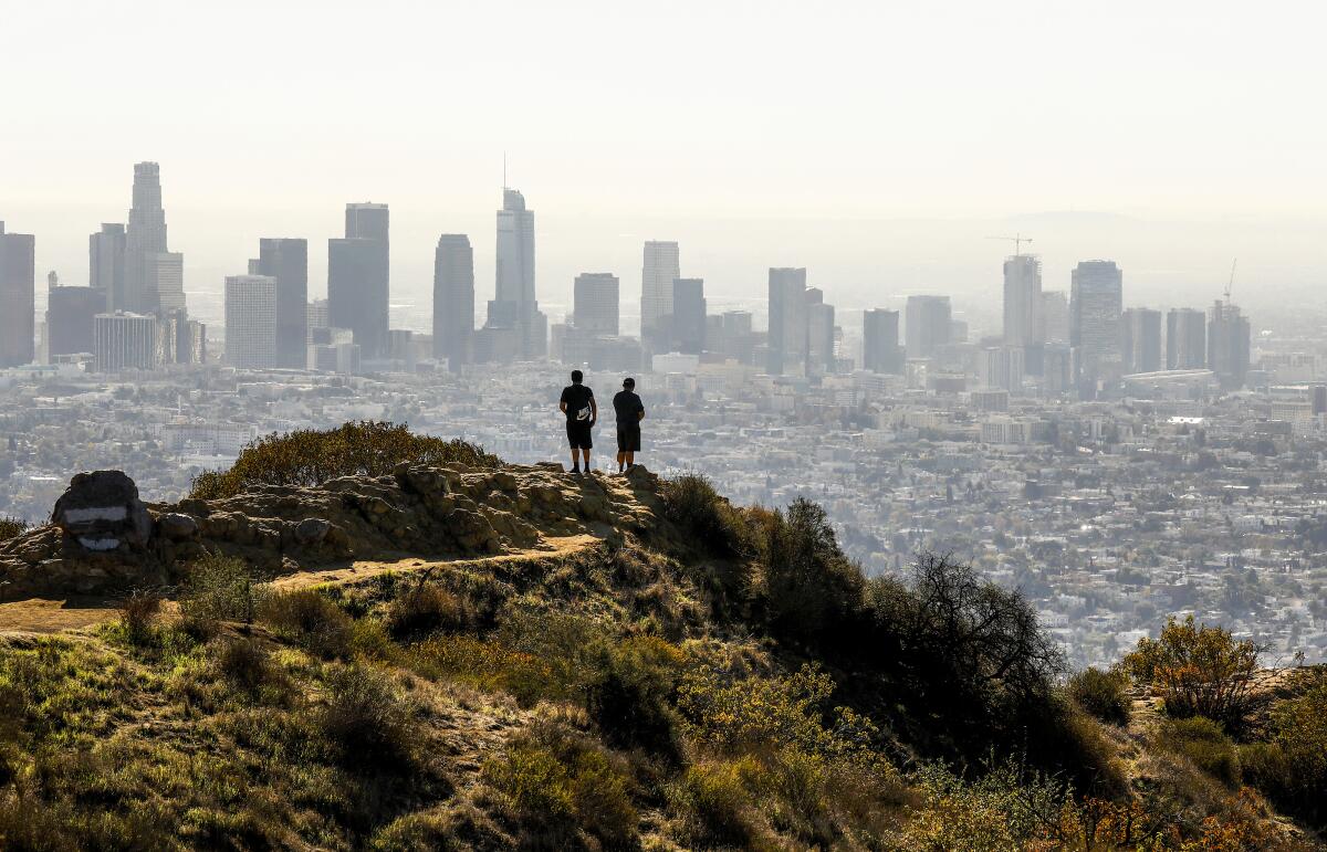 Two people stand on a brush-covered hill looking at neighborhoods and a city skyline in the distance.