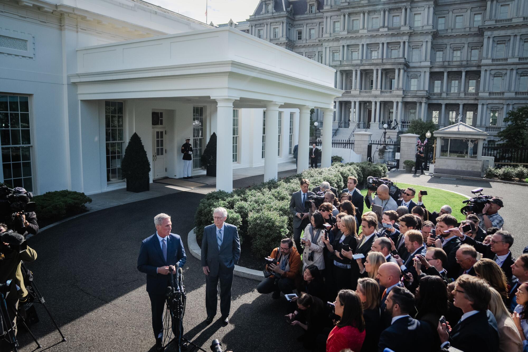 スーツを着た2人の男性がホワイトハウスの近くで他の数人の人々の前に立っています。