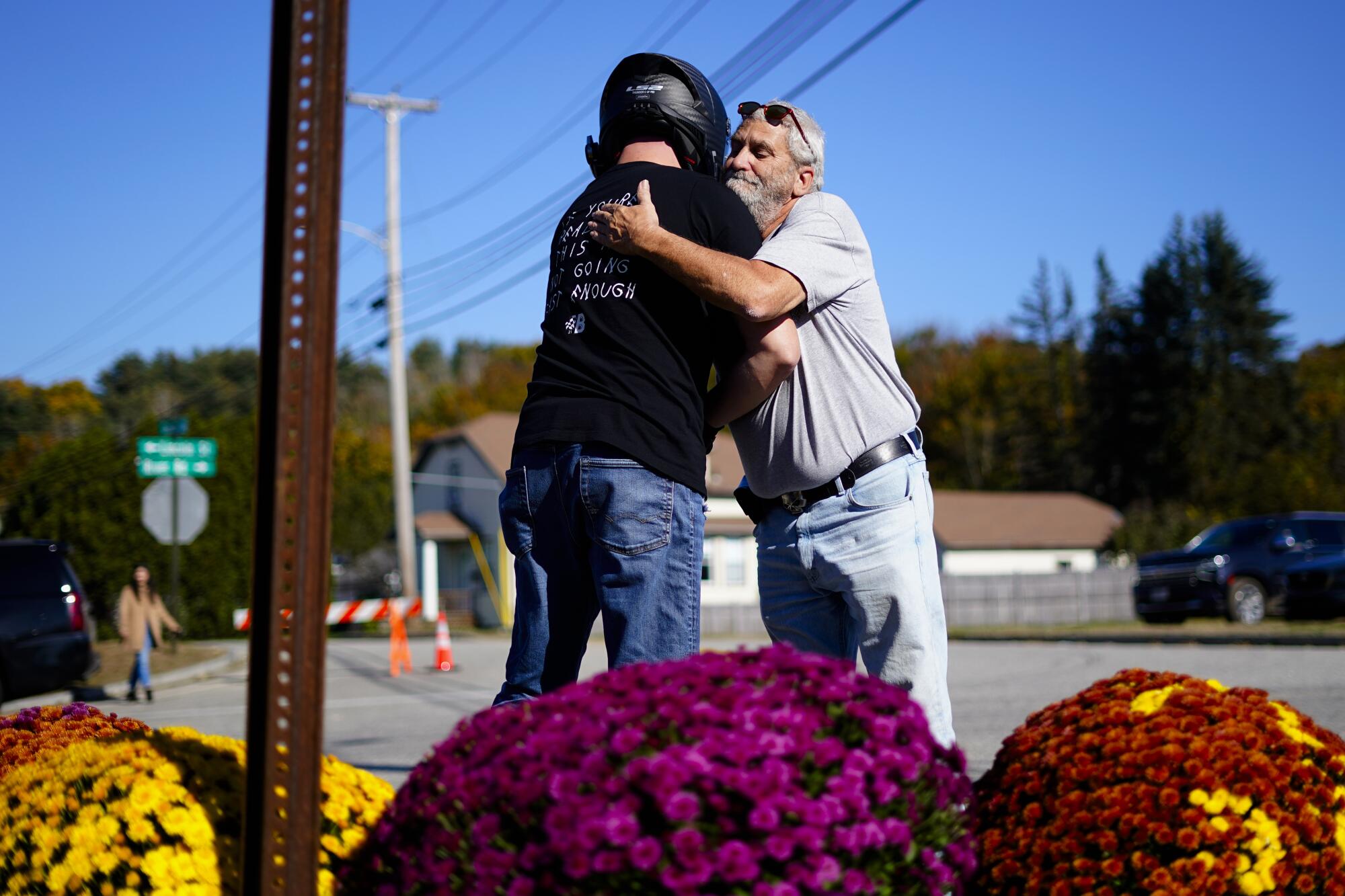 A man embraces a person in a motorcycle helmet next to bouquets of flowers