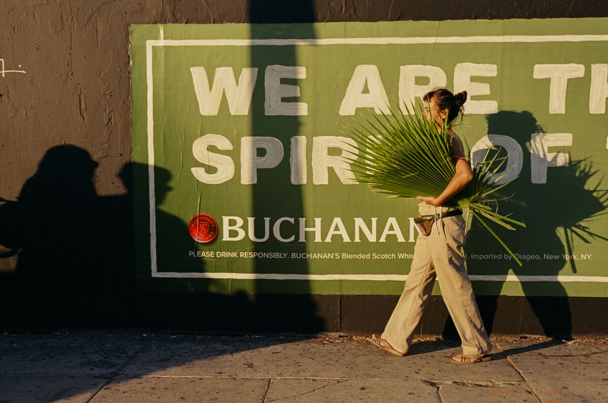 Artist Maria Maea, wearing a white tank and khakis, is seen carrying a bundle of palm fronds along a city sidewalk.