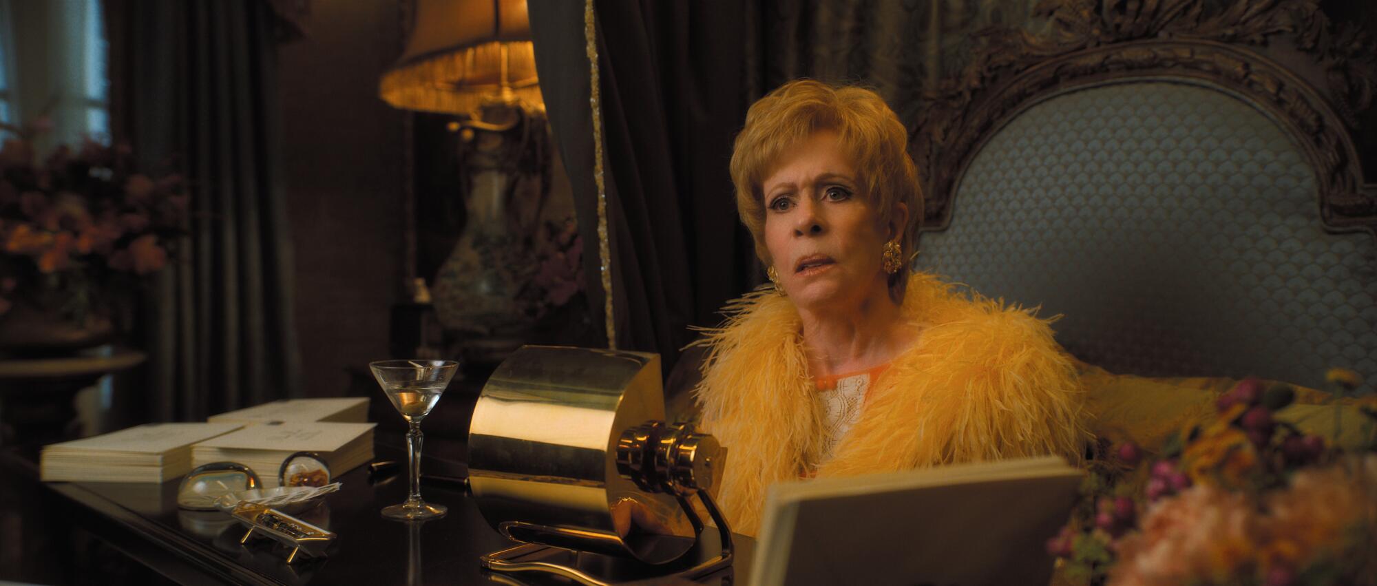 Carol Burnett wears a bright, feathery dressing gown while sitting at a desk in "Palm Royale."