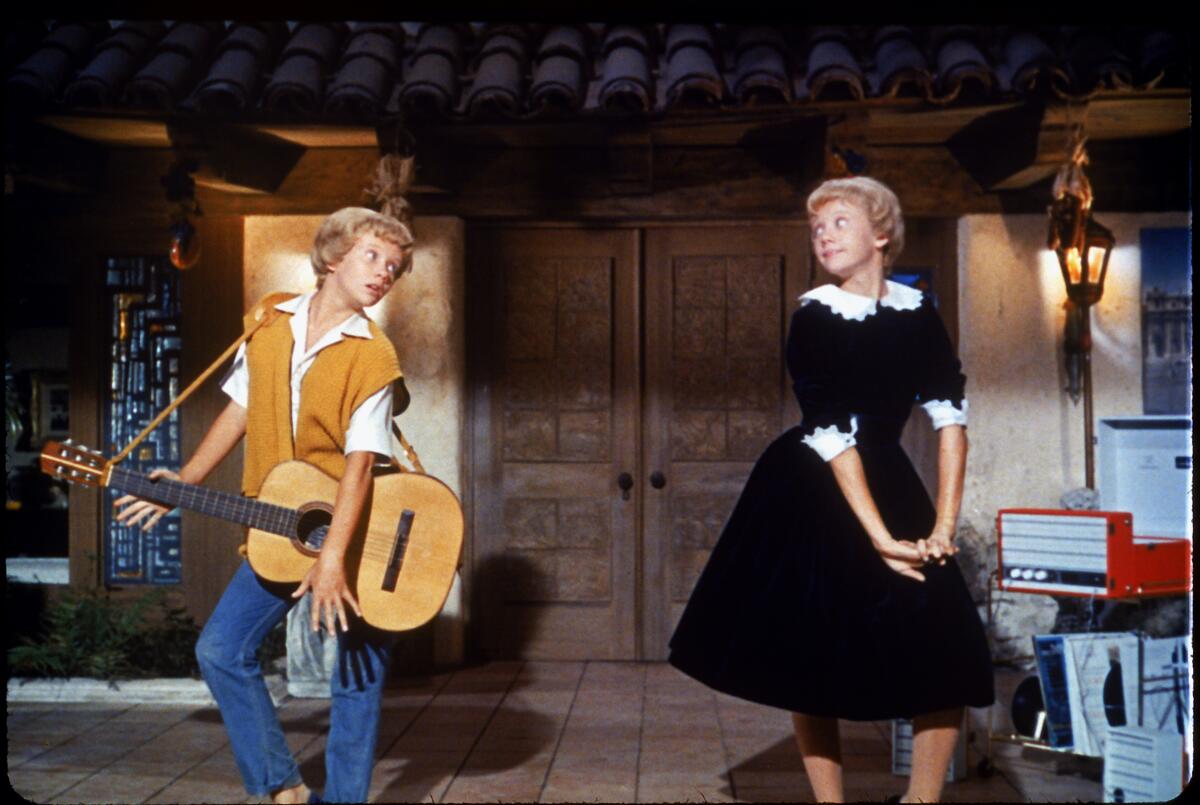 A girl in jeans holding a guitar, right, and an identical girl in a black dress. 