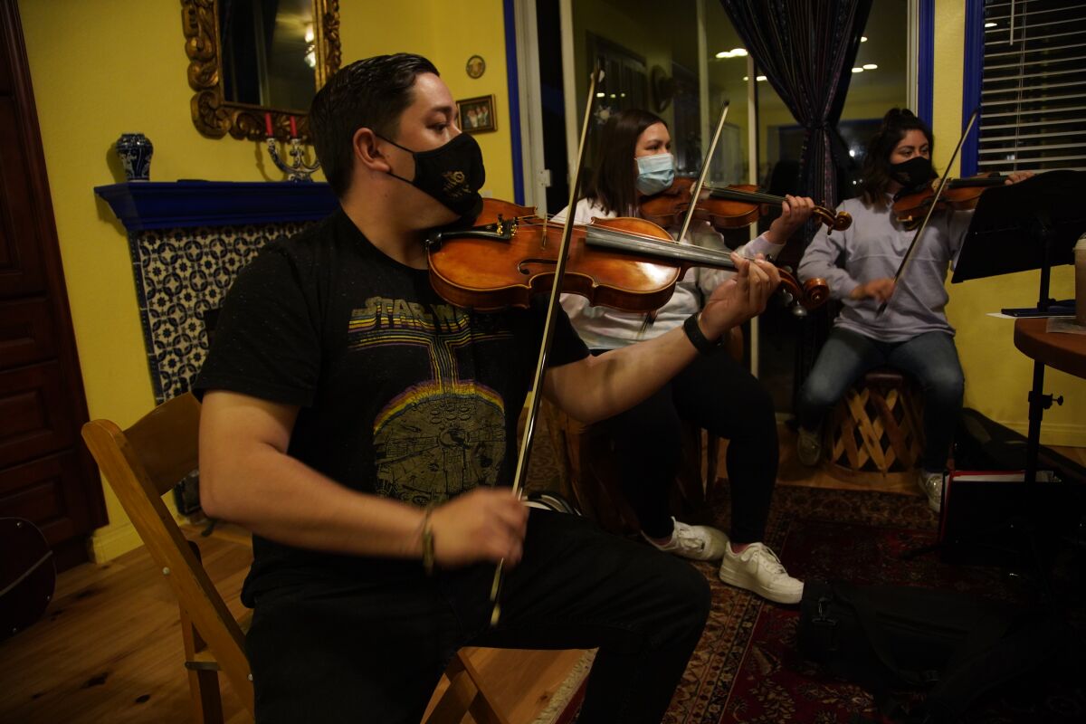 Three people, seated and wearing masks, play violins.