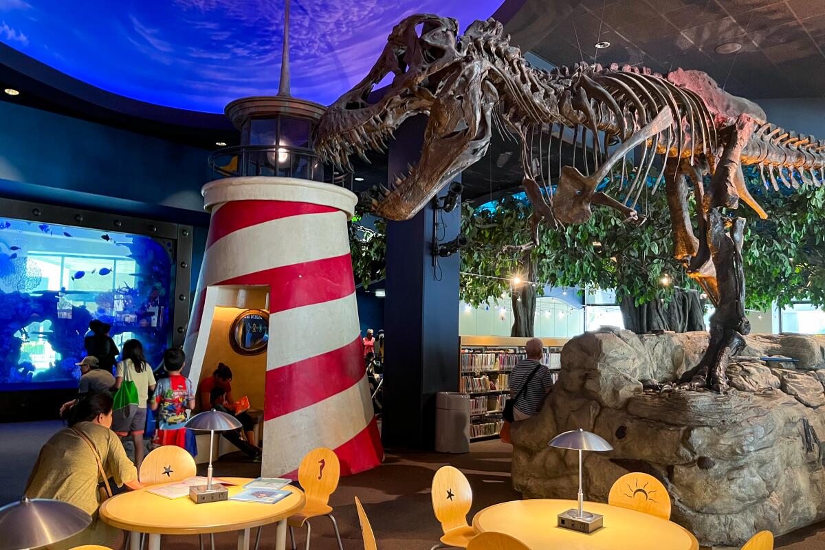 A T. rex skeleton looms over a red-and-white lighthouse and small round wood tables with chairs.