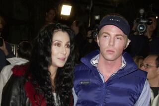 Cher in a black leather trench coat and Elijah Blue Allman are posing together in front of a group of photographers 
