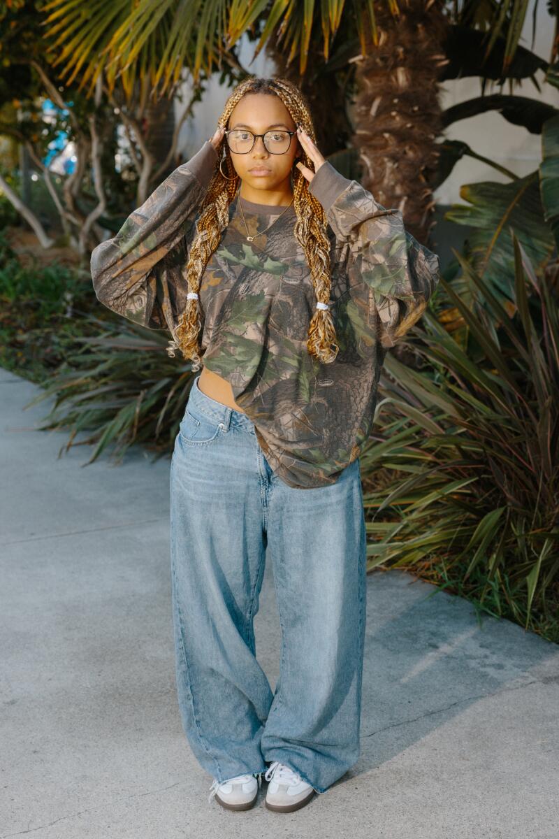 A girl posing for a portrait wearing glasses, a long-sleeved hunting camo shirt, baggy jeans and sneakers.