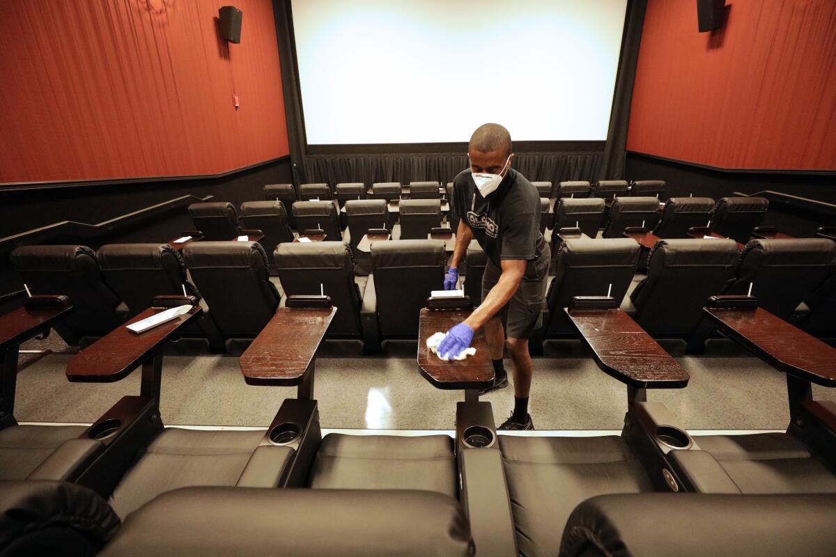 A man wearing a mask cleans the tables attached to movie seats in an empty theater.  