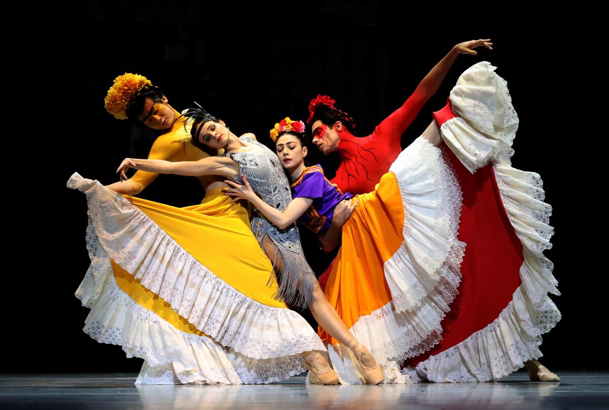 Three dancers in yellow, orange and red dresses collapse into another dancer in a shimmering silver dress.