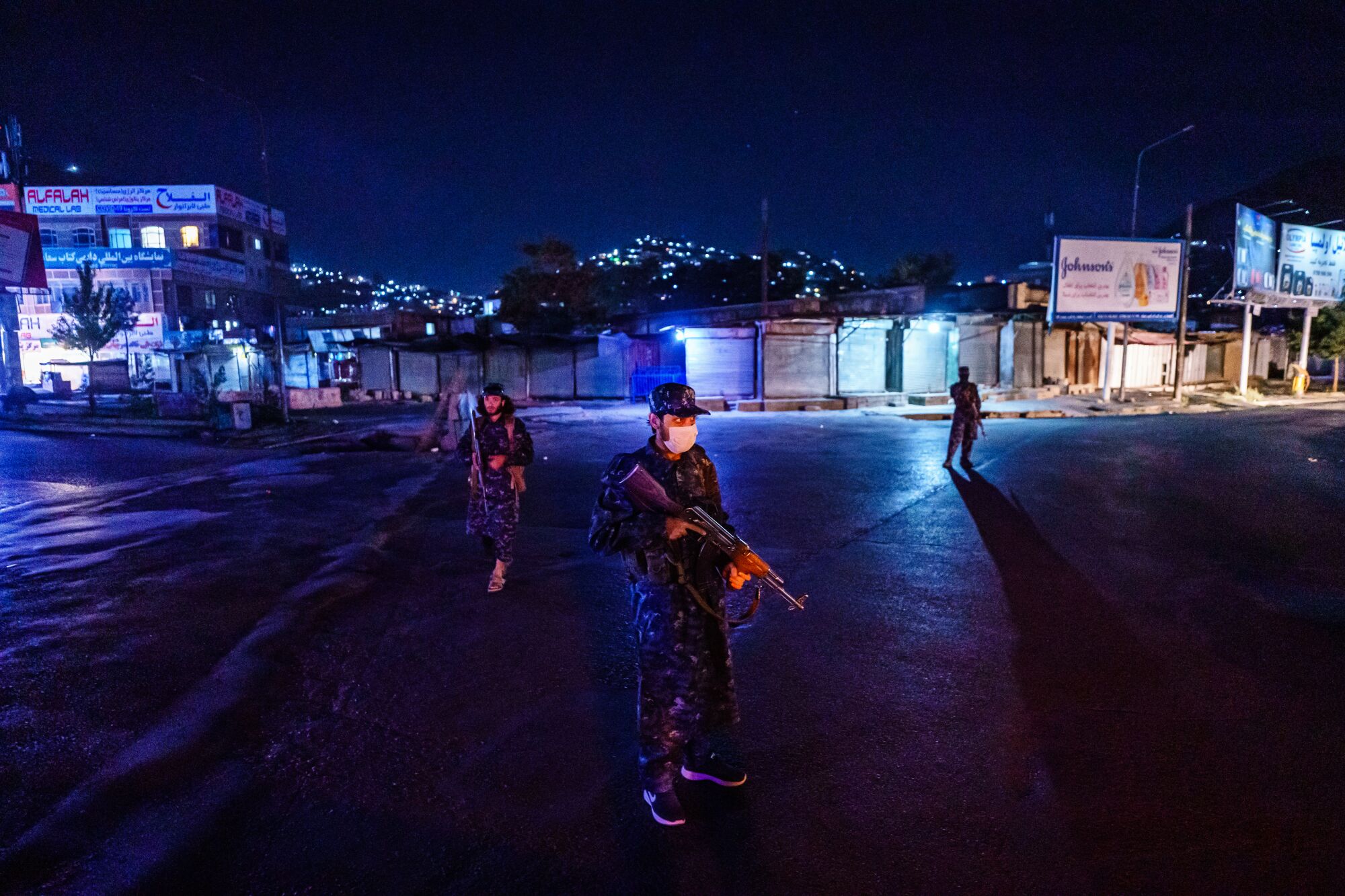 Men in fatigues stand holding their weapons in front of buildings with lights on at night.