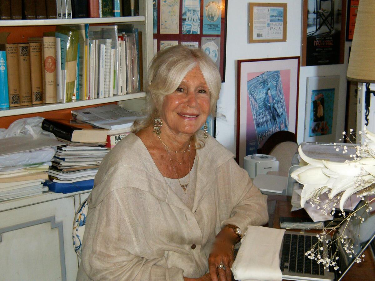 A white-haired woman smiles at her desk in a room filled with books and art