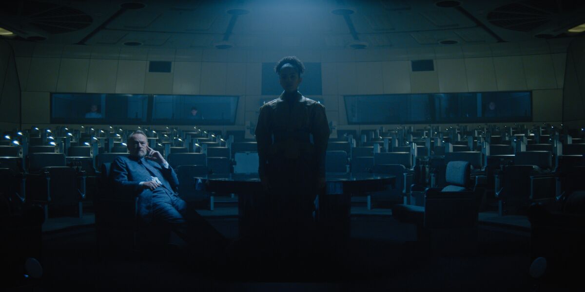 An illuminated man sits adjacent to a woman standing in shadows in an auditorium
