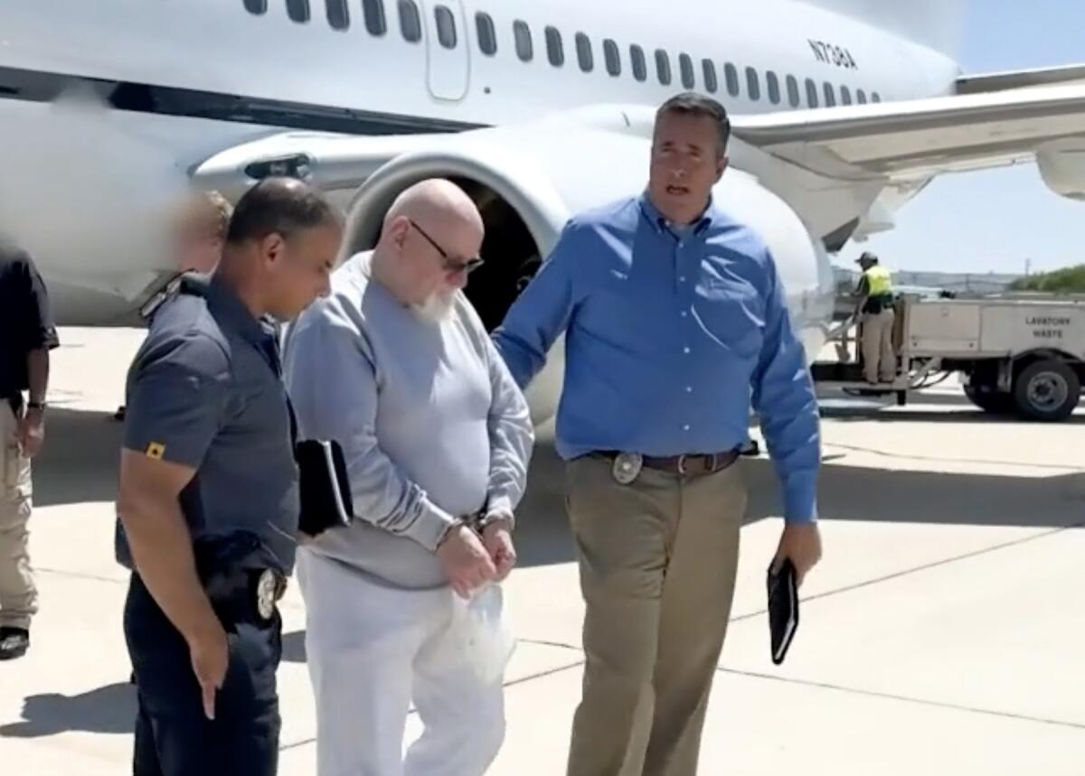 Warren Luther Alexander, with white goatee and bald head, walks with hands cuffed between two men on a tarmac.