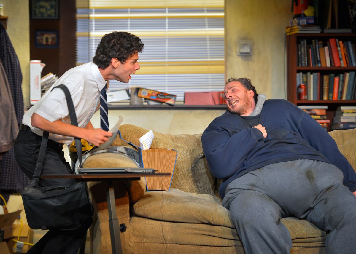 An actor in a shirt and tie talks to an obese man sitting on the couch at a play.