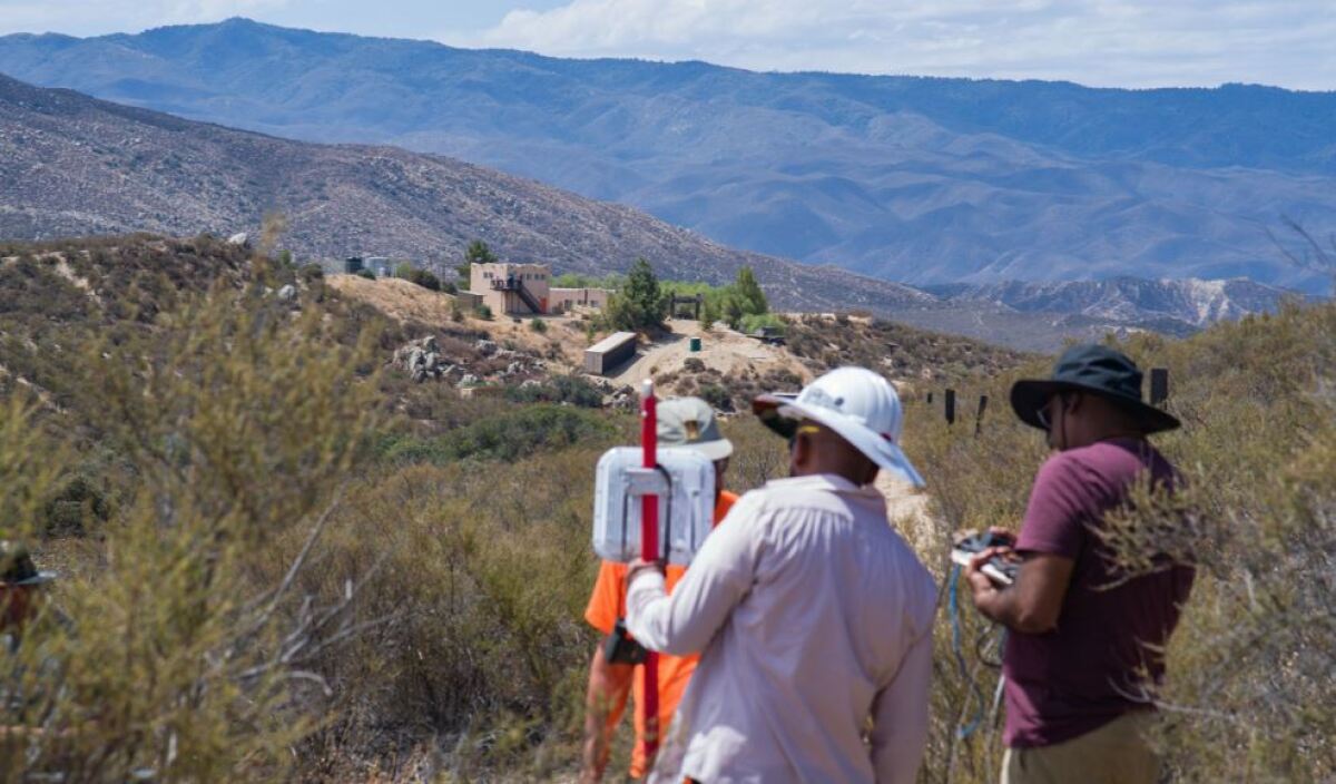 Two people wearing sun hats stand on a scrub-covered hillside installing radio equipment