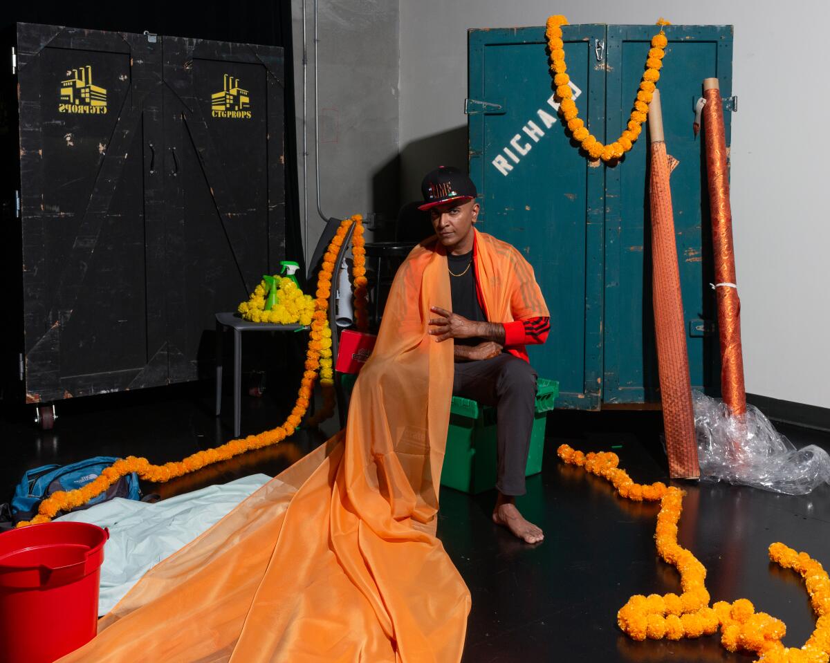 The comedian, actor and playwright D'Lo is seated and draped in orange cloth.