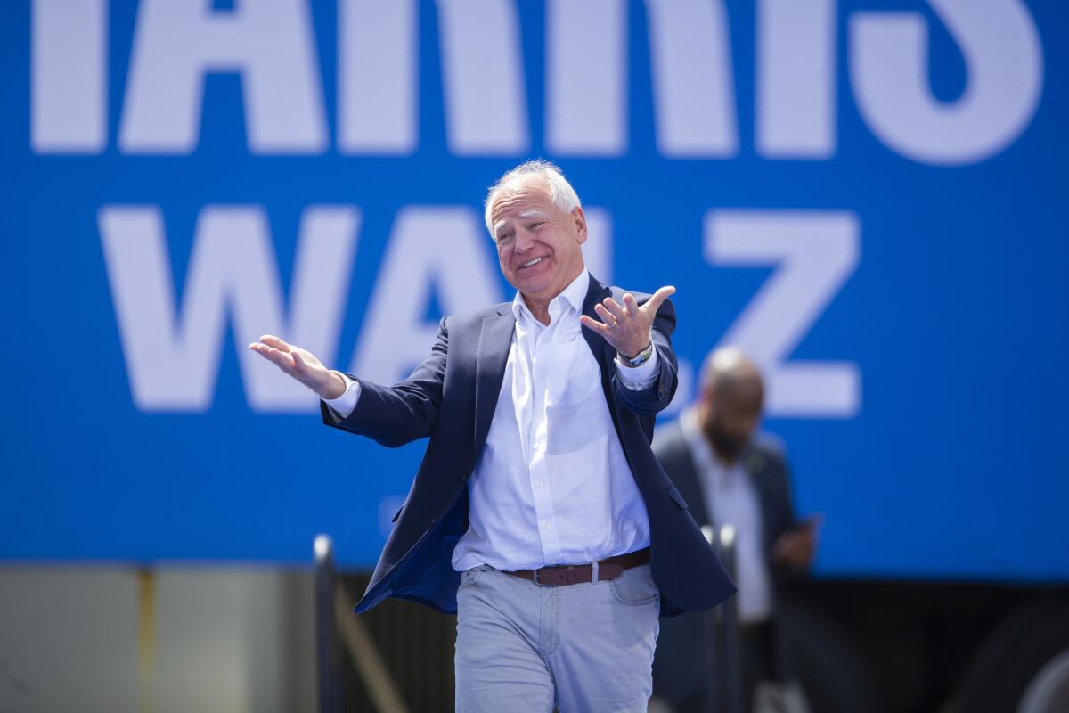 Tim Walz smiles and gestures during a campaign rally in front of a banner reading "Harris Walz."