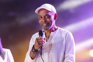 Frankie Beverly in white hat and linen shirt hold a microphone onstage