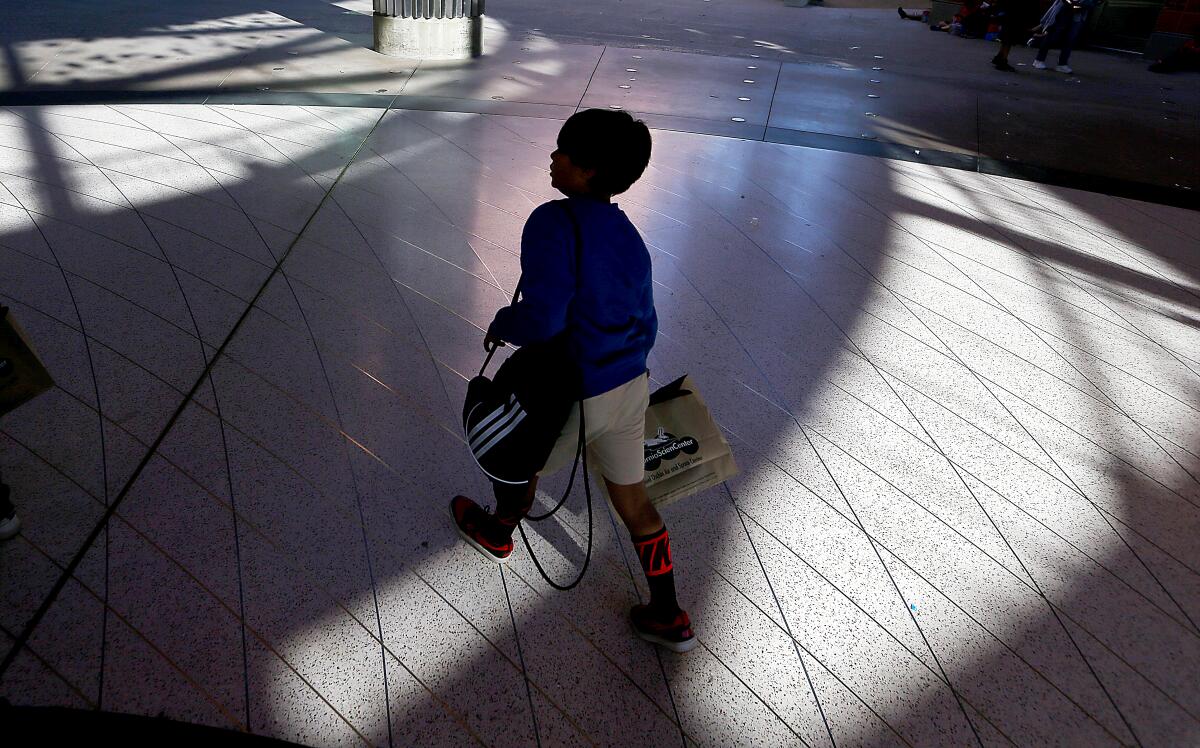 A child in sweat shirt and shorts and carrying a bag walks across a sun-dappled expanse of floor.