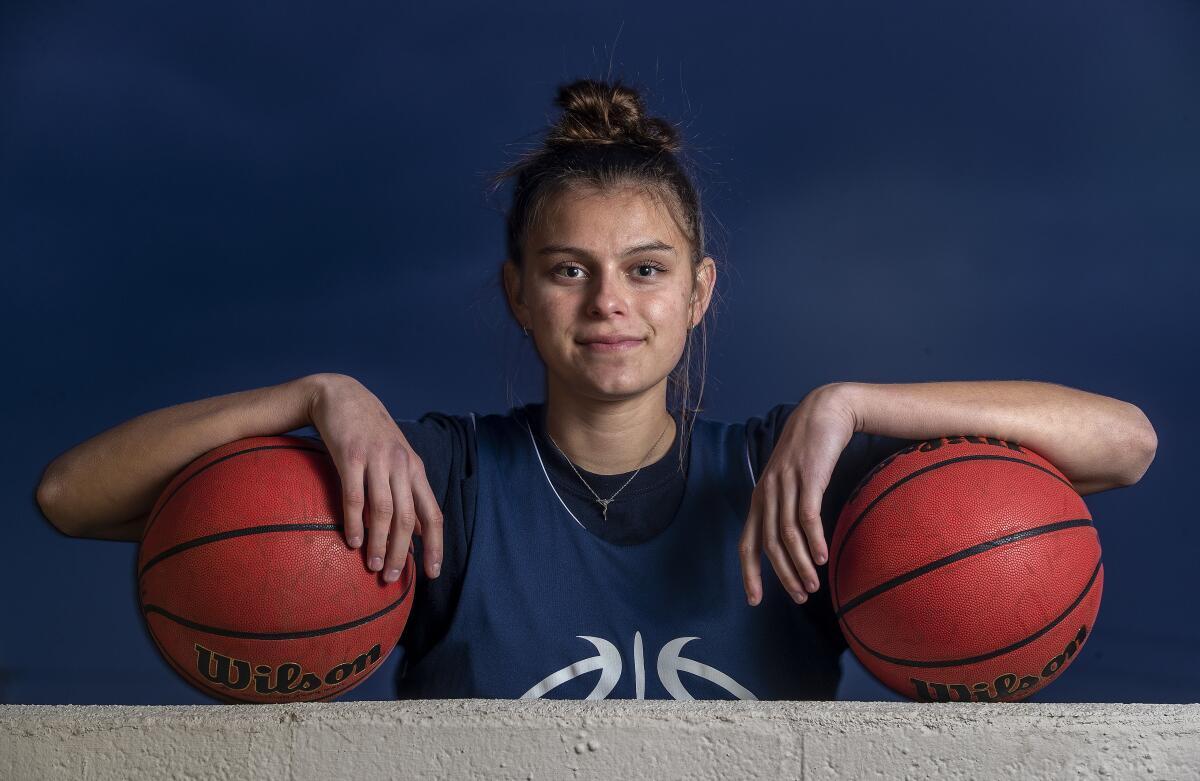 A girl poses with her arms draped over a pair of  basketballs on a ledge