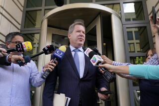 A man in dark suit and tie speaks as other people hold microphones toward him outside a building
