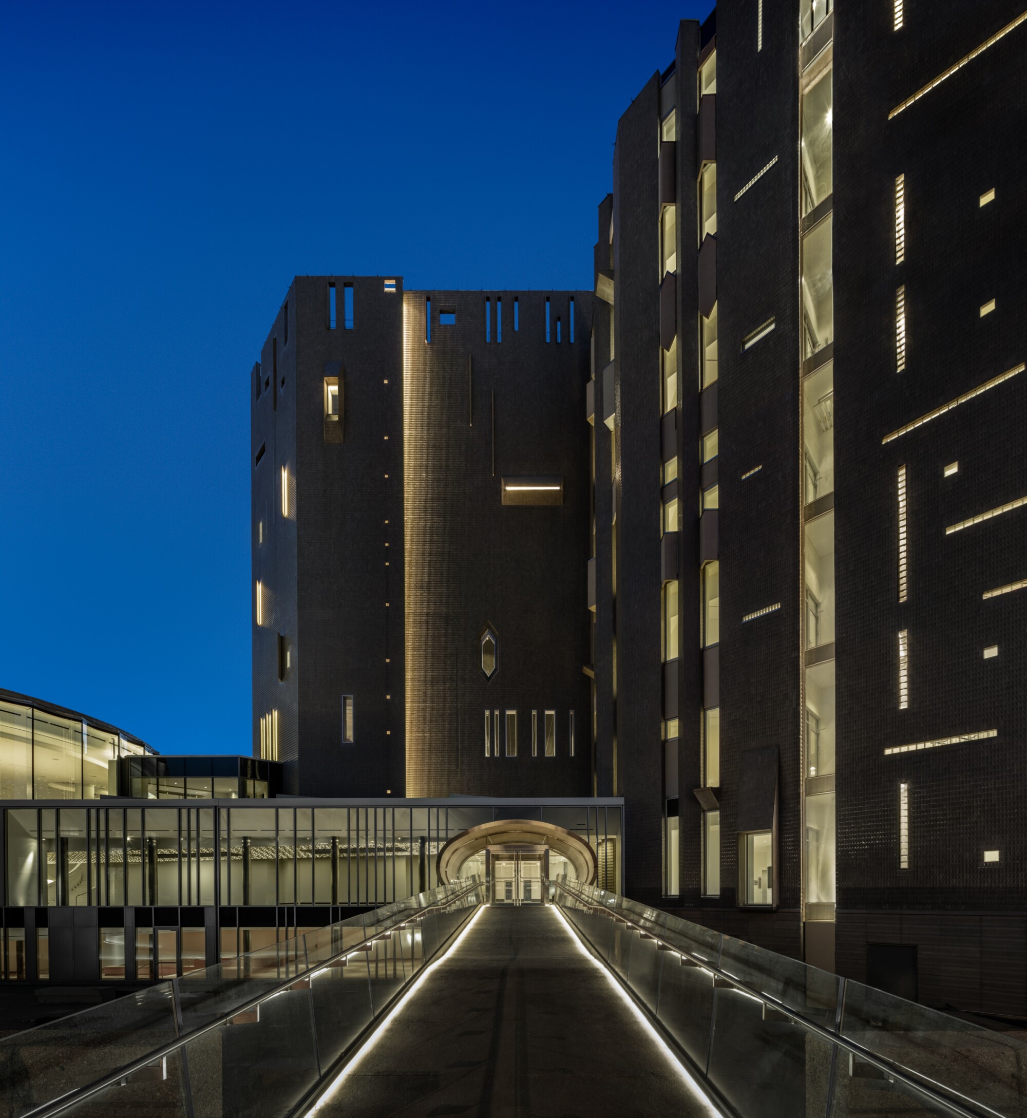 A long illuminated ramp leads to an oval doorway in a towering building illuminated from within and seen at dusk.