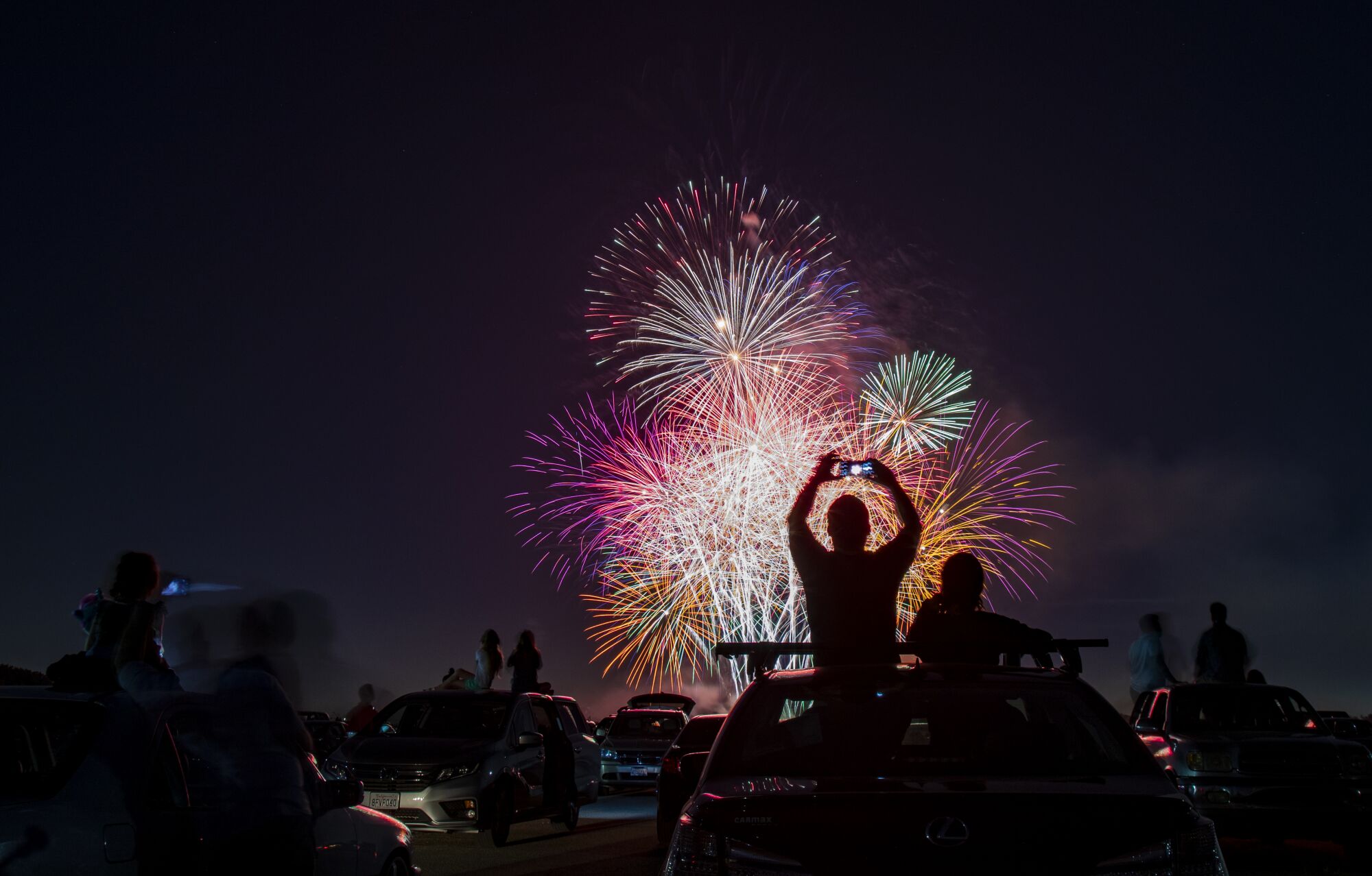 People sit atop their cars, holding up cellphones to take photos and video, as multiple fireworks explode. 