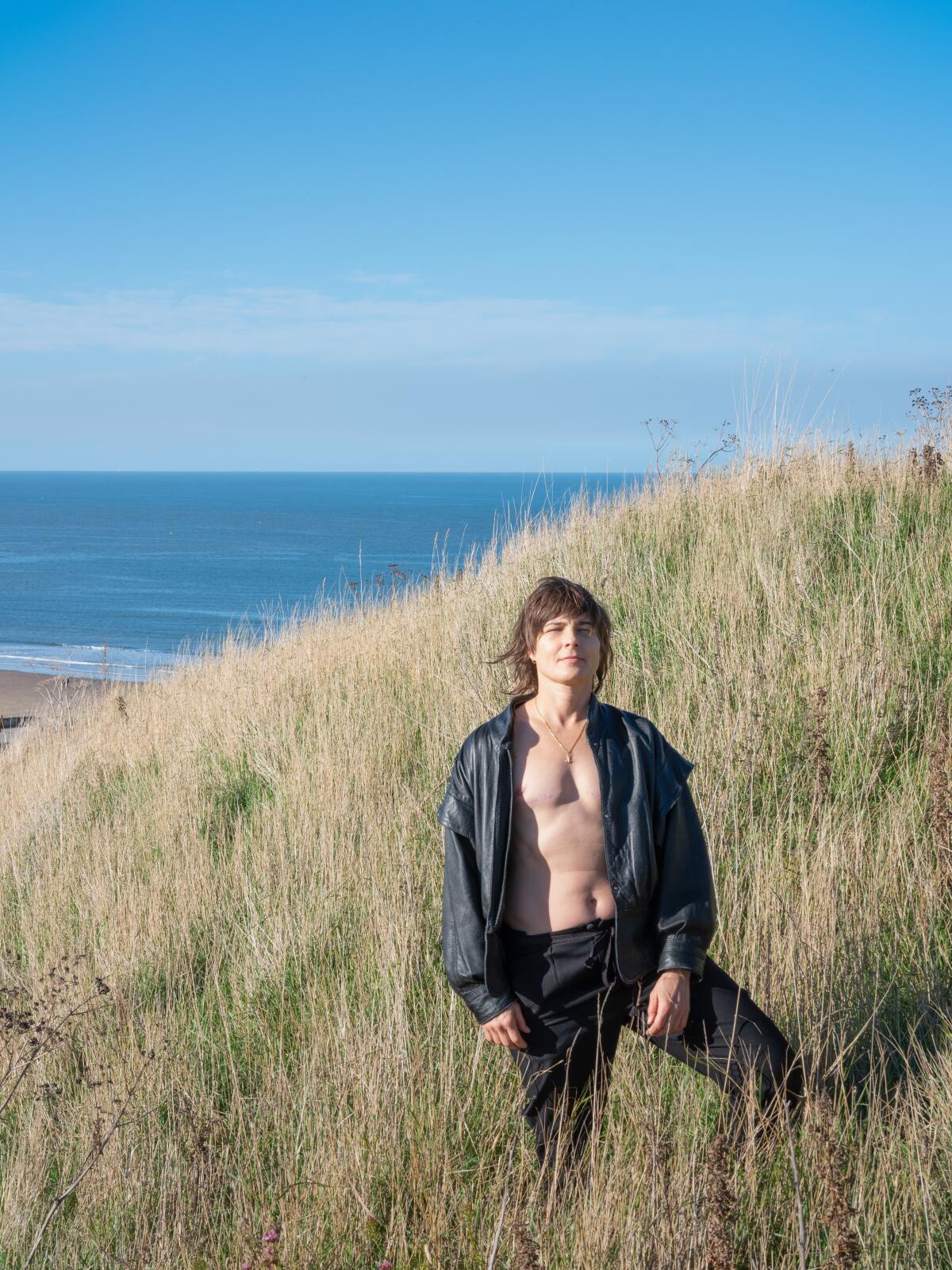 A nonbinary trans man stands in a field of sea grass in an open sweatshirt