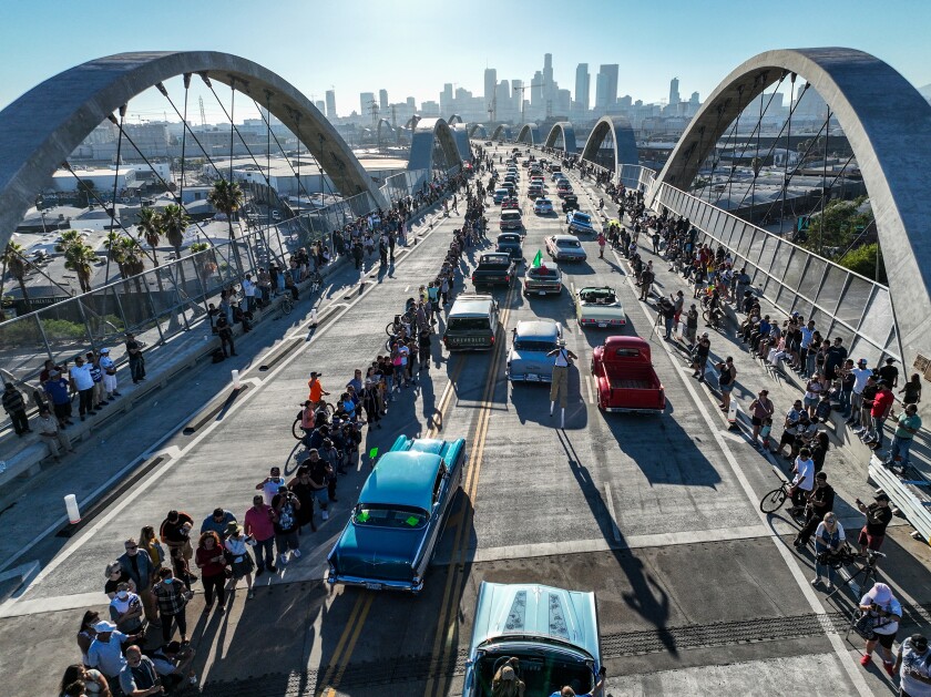 People line the sides of a bridge with many arches as a group of classic cars moves up the road.