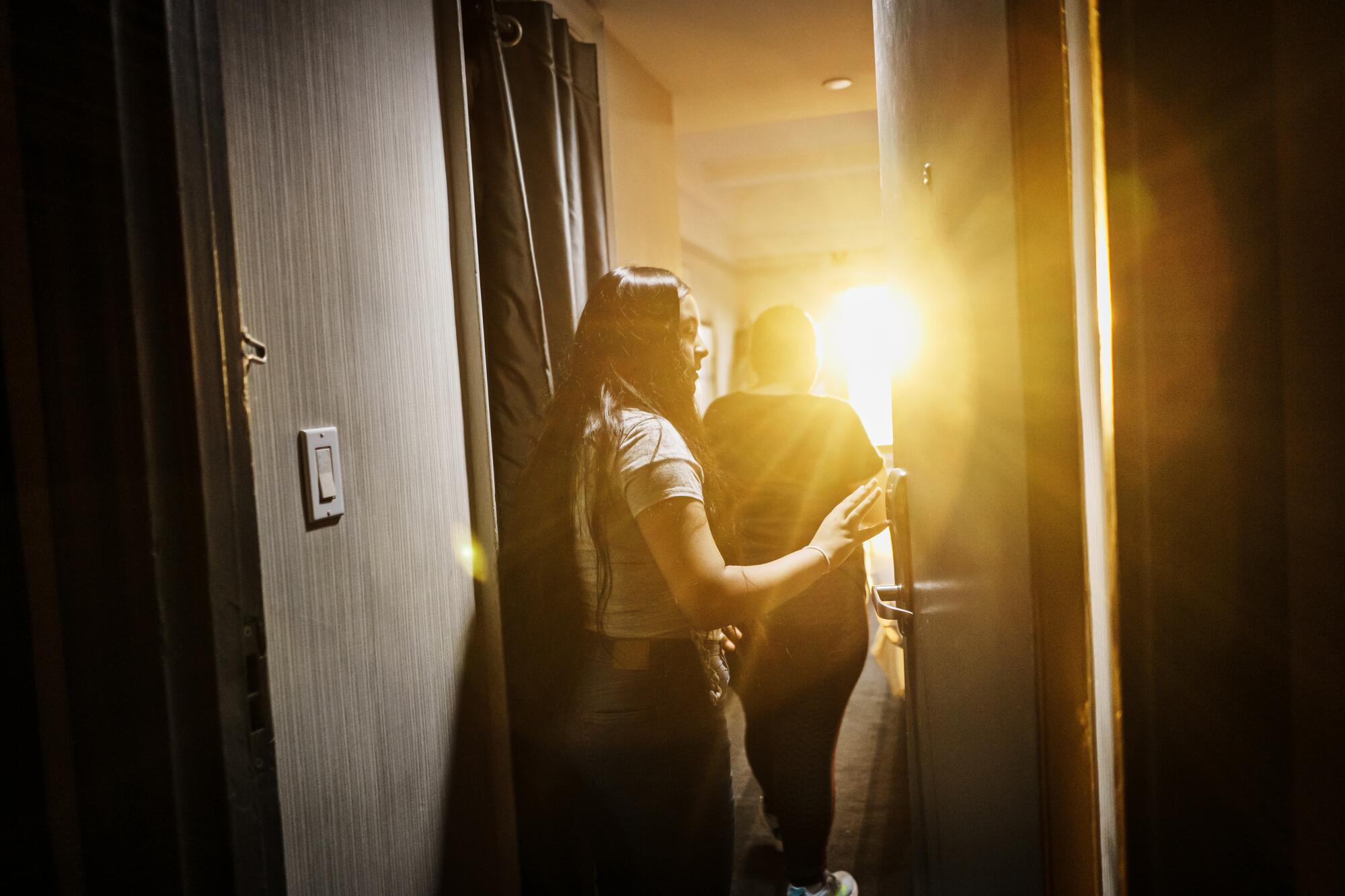 A girl and a woman enter a room as sunlight streams through a window. 