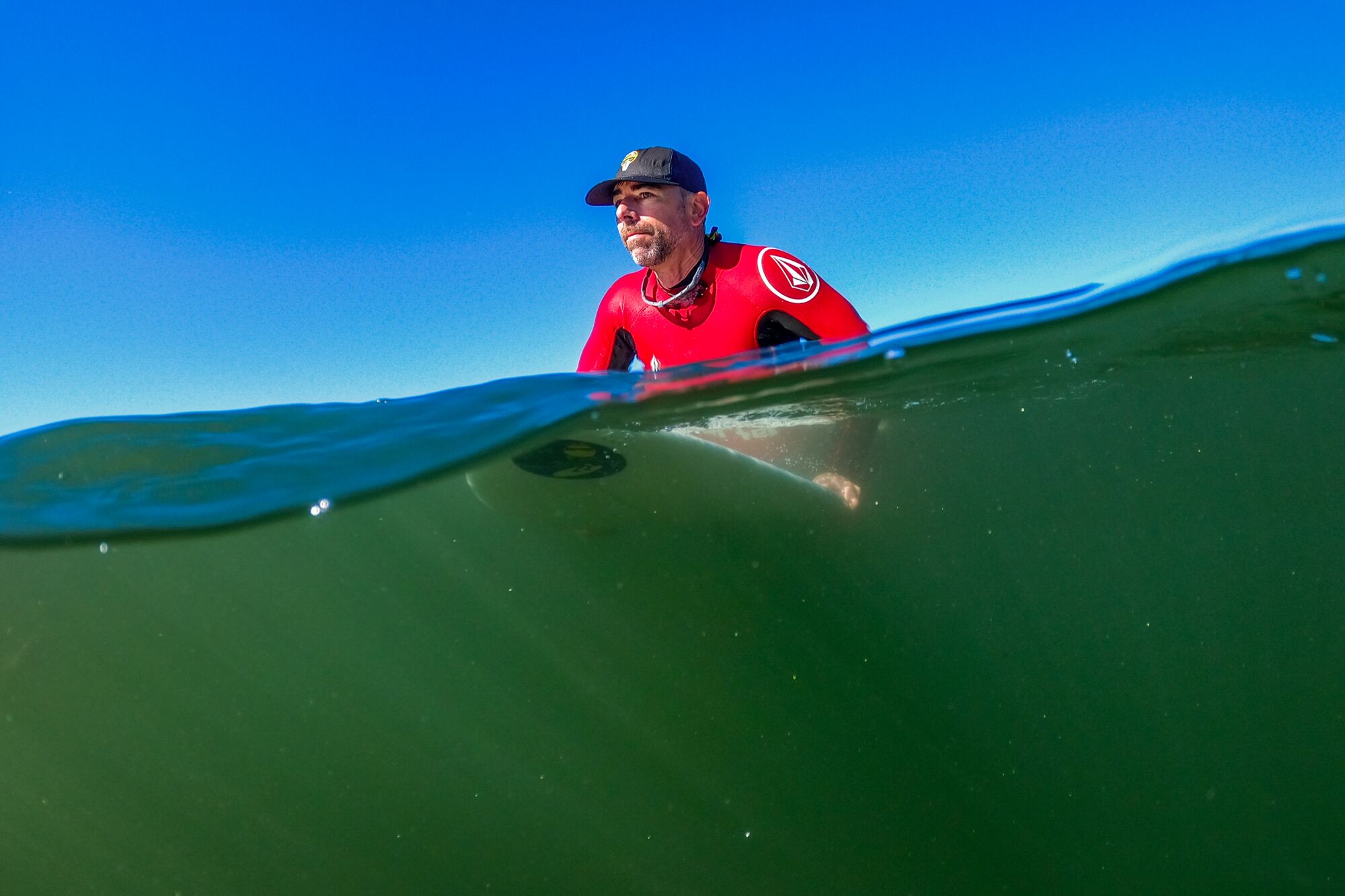 A man wearing a read and black wetsuit and a baseball cap sits on his surfboard in the water.