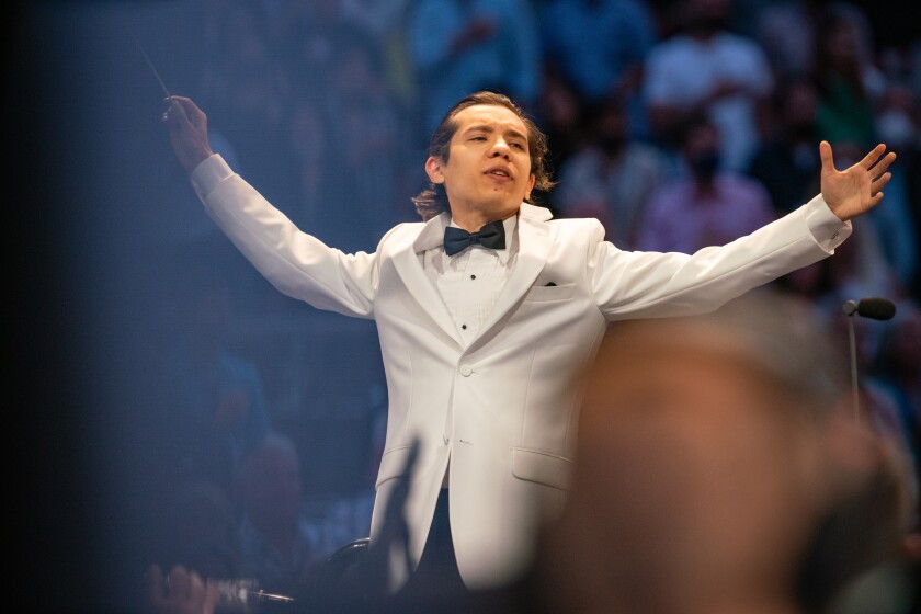 A conductor in a white suit jacket and bowtie spreads his arms wide